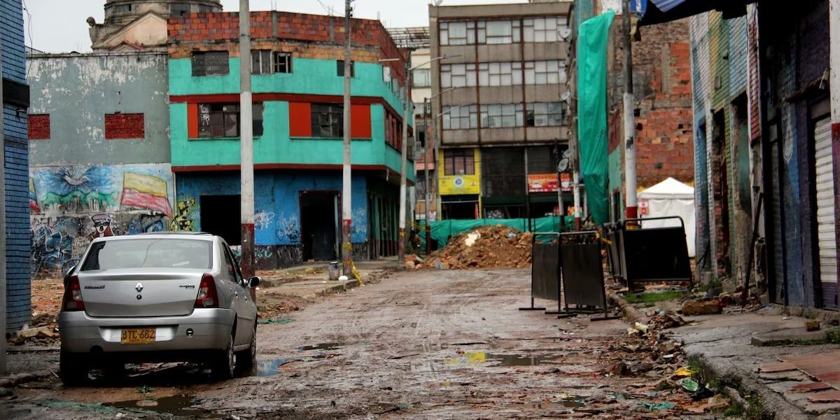 Calle sin pavimentar con charcos, un automóvil estacionado, edificios de ladrillo y concreto, escombros y vallas en la vía.