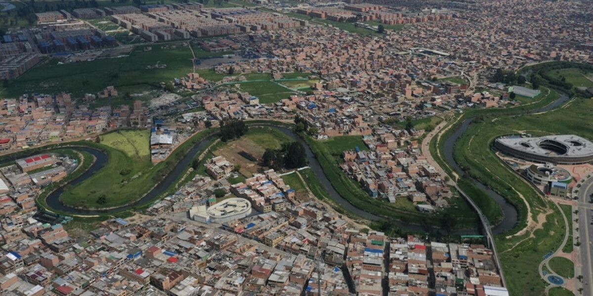 Vista aérea de una zona urbana con viviendas, áreas verdes, un río sinuoso, vías y un edificio circular a la derecha.