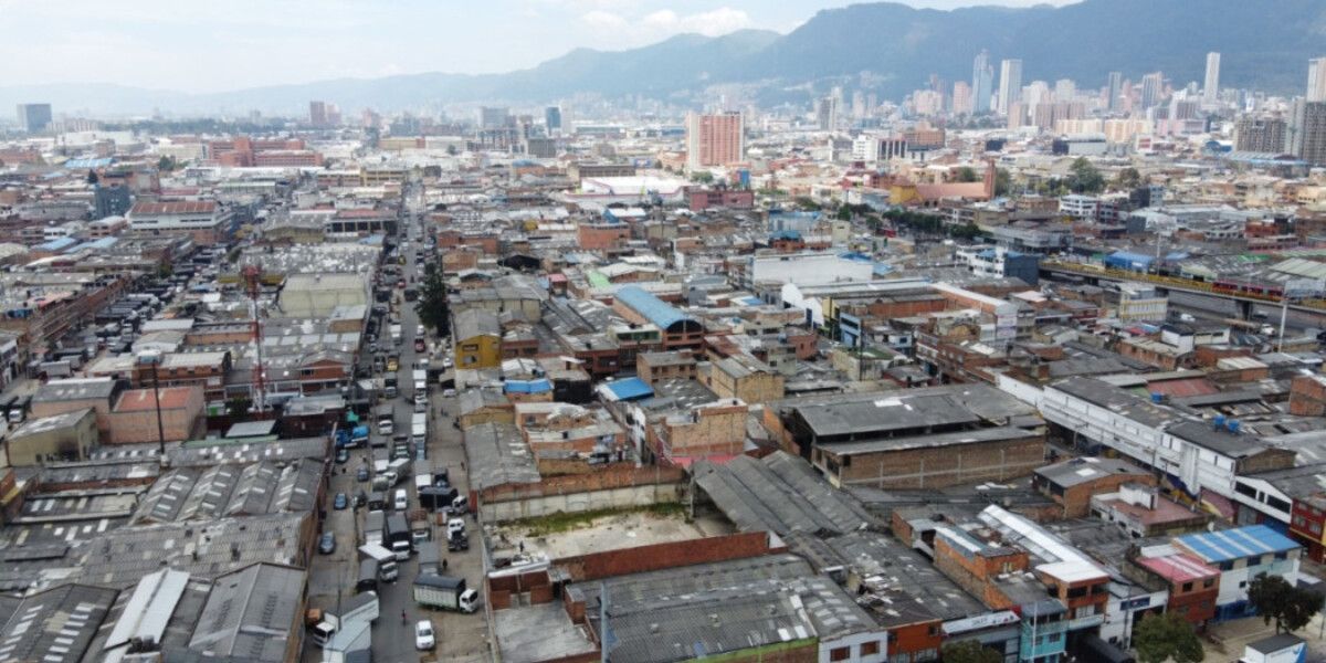 Vista aérea de un sector urbano con edificaciones de baja altura, una avenida con tráfico, edificios altos y montañas al fondo.