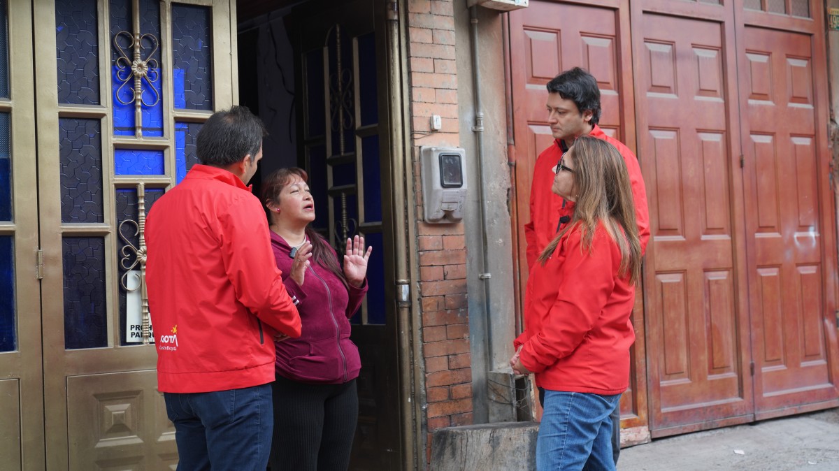 Tres personas con chaquetas rojas conversan con una mujer en la entrada de una vivienda.